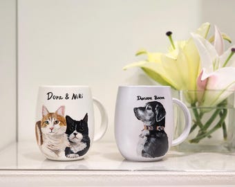 May include: Two white ceramic mugs with painted pet portraits. One mug features a cat portrait with the names "Dora & Niki". The other mug has a dog portrait with the name "Duncan Bacon". Both mugs have a curved shape and a white handle.