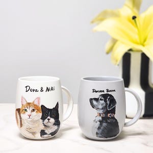 May include: Two white ceramic mugs with painted pet portraits. One mug features a cat with orange and white fur and a black and white cat, with the text "Dora & Niki". The other mug has a black and white dog with a collar, and the text "Duncan Bacon".