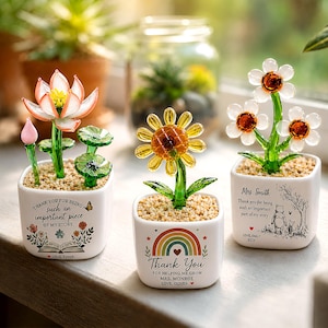 Op de afbeelding: Drie decoratieve glazen bloemstukken in witte vierkante potten. Eén heeft een lotusbloem, een andere een zonnebloem en de derde madeliefachtige bloemen. Elke pot heeft een bedankbericht en is gevuld met kleine kiezels.