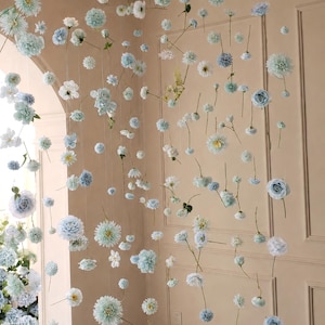 May include: A display of hanging artificial flowers in shades of white and blue. The flowers are suspended from the ceiling, creating a delicate and airy aesthetic. Various flower types are visible, including roses and daisies.