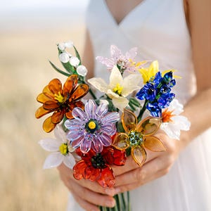 May include: A vibrant bouquet of glass flowers in shades of orange, red, purple, blue, yellow, and white. The flowers are held in a person's hands, with a blurred background of a field and sky. The bouquet includes various flower types.