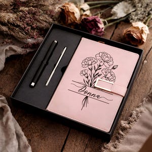 May include: A pink notebook with a floral design and the name "Donna" is displayed in a black gift box with a black pen and a silver pen. The notebook has a gold-colored clasp.