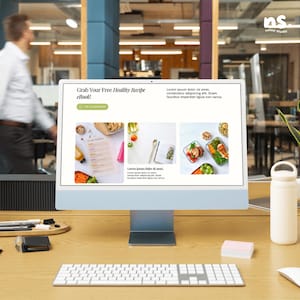 May include: A desktop computer displays a webpage with the text "Grab Your Free Healthy Recipe eBook!". The screen shows images of healthy recipes. A white keyboard, mouse, and water bottle are on the wooden desk. A person walks in the background.