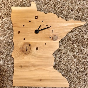 May include: A wooden clock shaped like the state of Minnesota with black hands and numbers. The clock is on a brown carpet.