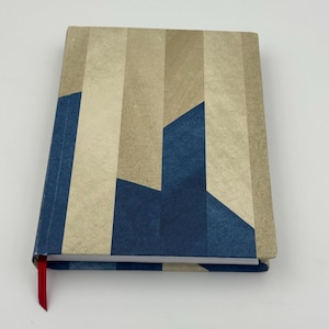 Può includere: Un taccuino chiuso con un motivo geometrico nei toni del beige e del blu. La copertina presenta strisce verticali e forme angolari. È visibile un segnalibro a nastro rosso. Il taccuino è su sfondo bianco.