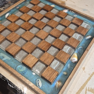 May include: A handcrafted wooden chess board with an ocean-themed design. The board has alternating squares of light wood and sand-coloured resin, with a blue resin border containing seashells. The board is set in a wooden frame.