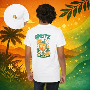 Peut inclure: T-shirt blanc avec un motif coloré "Spritz" au dos, représentant un cocktail à l'orange avec des glaçons, des tranches d'orange et des fleurs. Un petit motif orange et fleur est sur le devant. Le t-shirt est porté par une personne.