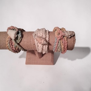 May include: Three bracelets made of crocheted yarn in muted pastel tones of pink, green, and cream. Each bracelet features a small, natural seashell attached to the woven cord, showcasing a textured, handmade appearance with soft color variations.
