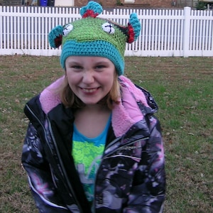 May include: A young person wearing a black and white patterned jacket with a pink fleece lining. They are wearing a green, blue, and red crocheted hat with a monster design.