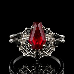 May include: A silver ring featuring a large, coffin-cut, red gemstone. The ring is embellished with black gemstones and a spiderweb design, giving it a gothic appearance. The ring is set against a black backdrop.
