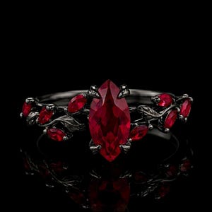 May include: A black metal ring featuring a large marquise-cut red gemstone at the center, flanked by smaller marquise-cut red stones and sculpted metal leaf details along the band, set against a solid black background with a clear reflection.