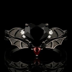 May include: A black metal ring featuring a large heart-shaped black center stone, flanked by two small clear stones on each side. The band is shaped like bat wings, with a small red heart-shaped stone set at the base of the central heart design.