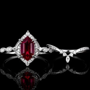 Peut inclure: Une parure de bagues en argent avec une pierre précieuse rouge foncé de taille hexagonale entourée d'un halo de petites pierres blanches. L'anneau présente un bord milgrain et des pierres blanches. Une alliance en V assortie est incluse.