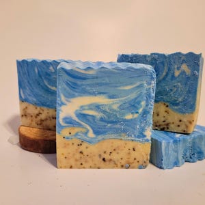 May include: Several bars of handmade soap with a beach theme. The soap features a blue and white swirled design on top, with a tan and speckled base. The edges of the soap are wavy, and a small wooden piece is visible.