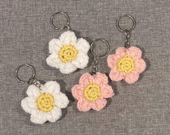 Könnte beinhalten: Vier handgefertigte Schlüsselanhänger mit gehäkelten Blumen. Zwei sind weiß mit gelben Zentren und zwei sind rosa mit gelben Zentren. Jede Blume hat einen silberfarbenen Schlüsselring.