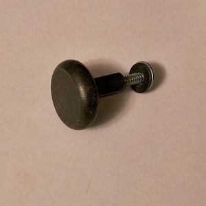 May include: A black cabinet knob with a round, flat head and a threaded screw. The knob is made of a dark, possibly painted material. The screw has a silver washer. The knob is likely used for furniture or cabinetry.