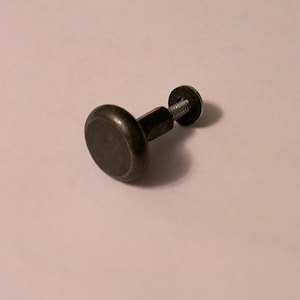 May include: A dark metal cabinet knob with a round, flat head and a threaded screw. The knob is a dark, muted color, likely bronze or a similar finish. The screw is attached to a smaller, circular piece.