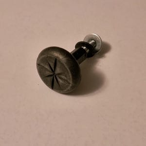 May include: A dark gray metal cabinet knob with a starburst design on the round head. The knob is attached to a black stem with a washer and screw. The hardware is likely for furniture or cabinetry.