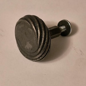 May include: A metal cabinet knob with a circular, ridged handle. The handle is attached to a hexagonal shaft and a screw with a washer. The knob is silver in color and appears to be made of steel.