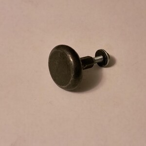 May include: A dark gray, round cabinet knob with a threaded post and a silver-colored washer and screw. The knob has a slightly textured surface and is designed for furniture or cabinetry.