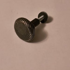May include: A dark gray, metal cabinet knob with a ridged, rounded face. The knob is attached to a threaded bolt with a small, round nut. The hardware is likely for a drawer or cabinet door.