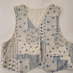 Könnte beinhalten: Eine ärmellose Weste mit einem Patchwork-Design aus cremefarbenem und blauem Stoff mit verschiedenen Blumen- und Streifenmustern. Das Kleidungsstück hat einen V-Ausschnitt, drei Knöpfe auf der Vorderseite und zwei untere Taschen aus Cord.