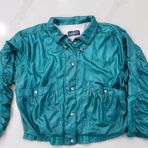 May include: A teal windbreaker jacket featuring a high collar with a drawstring, a front snap-button closure, and two front pockets. The jacket has long sleeves with gathered detailing and a white interior lining, with a Top Seed brand label.