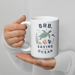 Puede incluir: Una taza de cerámica blanca con un gráfico de una tortuga marina nadando entre corales y algas. El texto dice BRB, SAVING THE OCEAN en letras azul oscuro. El diseño incluye pequeñas burbujas y detalles de peces en tonos azules y naranjas.