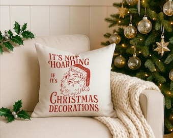 Puede incluir: Un cojín decorativo cuadrado de color crema con un diseño de Papá Noel rojo y el texto "It's Not Hoarding If It's Christmas Decorations." El cojín está sobre un sofá beige, cerca de un árbol de Navidad.