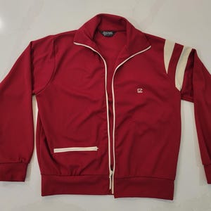 May include: A red track jacket featuring a full-length front zipper, a high collar, and long sleeves with ribbed cuffs. The jacket includes cream-colored accents on the shoulders and a small cream-colored logo on the chest, plus a zippered pocket.