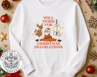 Könnte beinhalten: Weißes Sweatshirt mit einem Retro-Grafikdesign von Weihnachtsmann, Rentier und Schneemann, mit dem Text "Will Work For Christmas Decorations" in Orange. Das Design hat eine Vintage-Ästhetik, ideal für die Weihnachtszeit.