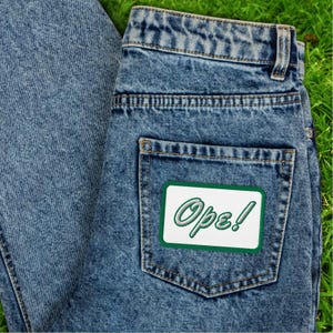 May include: Close-up of blue denim jeans with a white and green embroidered patch on the back pocket. The patch reads "Ope!" in a cursive font with a green border. The jeans are on a green surface.
