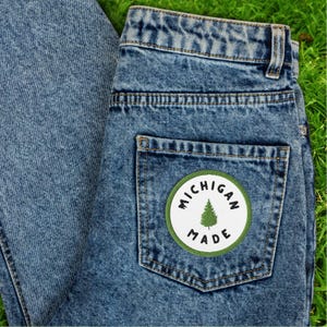 May include: Light blue denim jeans with a patch on the back pocket. The patch is a white circle with the words "MICHIGAN MADE" around a green pine tree graphic. The jeans are on a green surface.