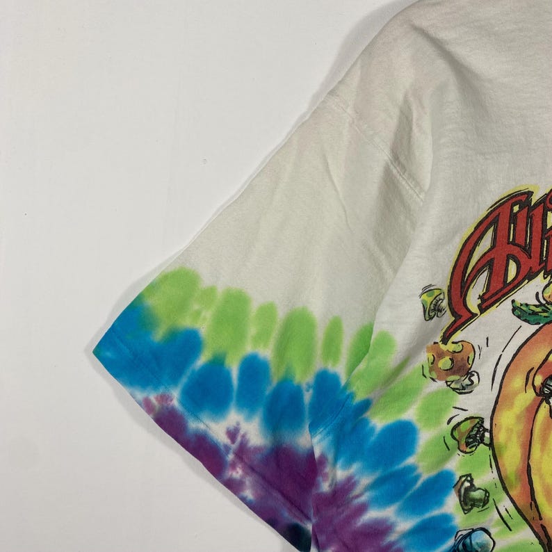 May include: White t-shirt with a tie-dye sleeve in shades of blue, green, and purple. The shirt features a colorful graphic design on the front. The sleeve is a tie-dye pattern.