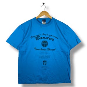 May include: A bright blue t-shirt with the text "Defense Organization Border" and "Tamakoma Branch" in black. The shirt has a crew neck and short sleeves, hanging on a wooden hanger.