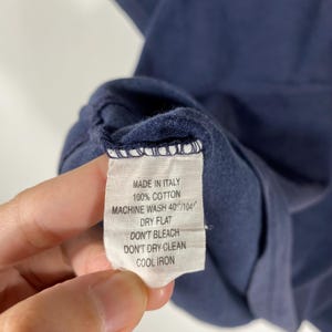 May include: A close-up of a white clothing tag with black text. The tag reads "MADE IN ITALY", "100% COTTON", "MACHINE WASH 40&deg;/104&deg;", "DRY FLAT", "DON'T BLEACH", "DON'T DRY CLEAN", and "COOL IRON". The tag is attached to a navy blue garment.