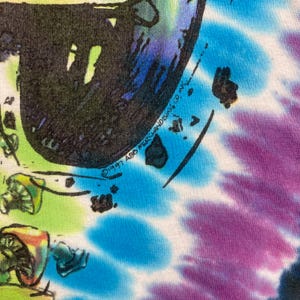 May include: Close-up of a tie-dye t-shirt with a colorful design. The shirt features a black graphic with a mushroom and other psychedelic imagery. The background is a mix of blue, purple, green, and yellow. The text "&copy;1997 ABG MERCHANDISING CO INC" is visible.