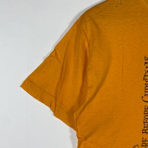 Puede incluir: Camiseta de color amarillo mostaza con detalles desgastados. El lado derecho de la camiseta presenta texto negro de una pel&iacute;cula. La manga corta es visible. La camiseta est&aacute; hecha de un material suave.