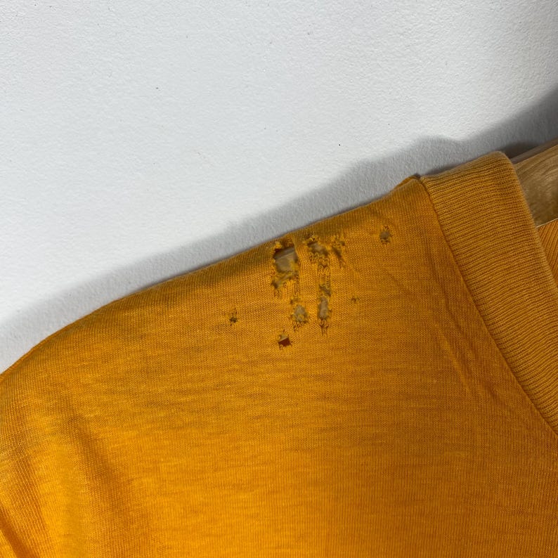 Puede incluir: Camiseta amarilla mostaza con cuello en V y detalles desgastados. La zona del hombro de la camiseta muestra m&uacute;ltiples agujeros y signos de desgaste. La camiseta est&aacute; colgada en una percha de madera sobre un fondo blanco.