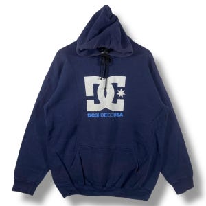 May include: Navy blue hoodie with a white DC Shoes logo and a star graphic. The text "DCSHOECOUSA" is printed below the logo in blue. The hoodie has a drawstring hood and a front pocket.
