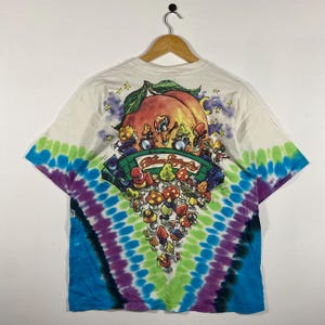 May include: White t-shirt with a tie-dye pattern in blue, green, and purple. The back of the shirt features a graphic of mushrooms playing instruments in front of a peach. The text "Allman Brothers &" is printed on the graphic.