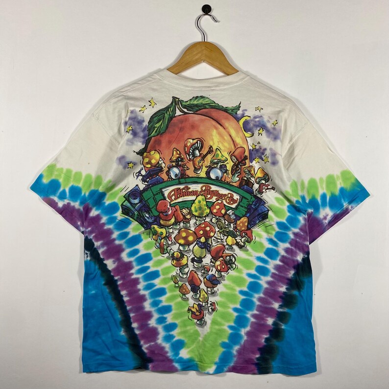 May include: White t-shirt with a tie-dye pattern in blue, green, and purple. The back of the shirt features a graphic of mushrooms playing instruments in front of a peach. The text "Allman Brothers &" is printed on the graphic.