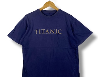 May include: Navy blue t-shirt with the word "TITANIC" printed in gold across the chest. The shirt is hanging on a wooden hanger against a white background. The t-shirt has short sleeves and a crew neck.