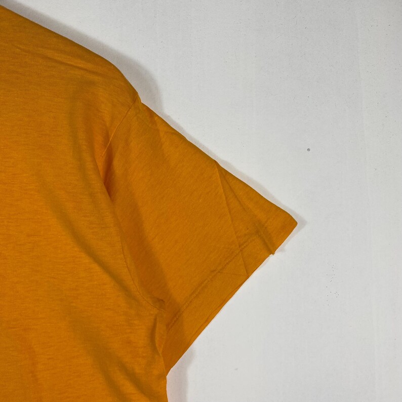 Puede incluir: Primer plano de una secci&oacute;n de una camiseta de color amarillo mostaza. La tela tiene un aspecto ligeramente texturizado. La manga y el hombro son visibles sobre un fondo blanco. Las costuras est&aacute;n claramente cosidas.