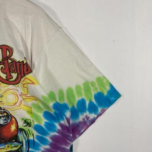 May include: White t-shirt with a colorful graphic design and tie-dye sleeves. The graphic features the text "The Band" in red and yellow, with a cartoon image of a vehicle. The sleeves are tie-dyed in shades of green, blue, and purple.