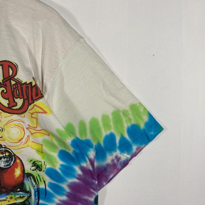May include: White t-shirt with a colorful graphic design and tie-dye sleeves. The graphic features the text "The Band" in red and yellow, with a cartoon image of a vehicle. The sleeves are tie-dyed in shades of green, blue, and purple.