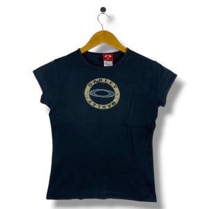 May include: A dark navy blue short-sleeve t-shirt featuring a circular graphic on the chest. The graphic contains the word OAKLEY in a distressed font surrounding a central oval logo. The shirt has a crew neckline and a red brand label inside.