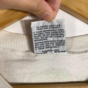 May include: Close-up of a white garment tag with care instructions printed in black. The tag reads "TO INSURE LONG LIFE FROM THIS GARMENT" and includes washing and drying instructions. The tag also states "MADE IN USA".