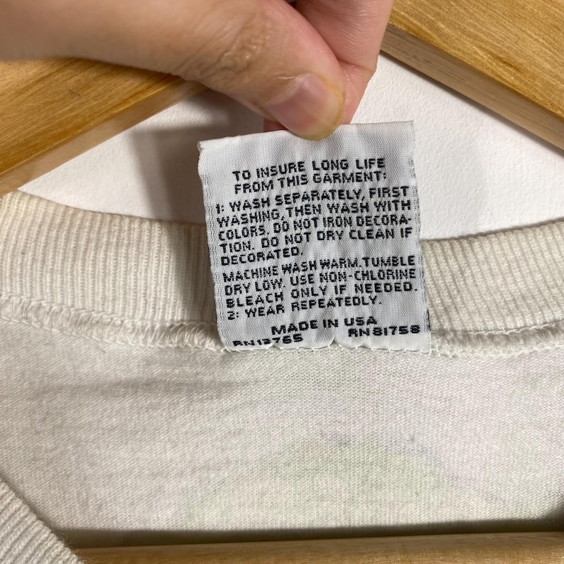 May include: Close-up of a white garment tag with care instructions printed in black. The tag reads "TO INSURE LONG LIFE FROM THIS GARMENT" and includes washing and drying instructions. The tag also states "MADE IN USA".