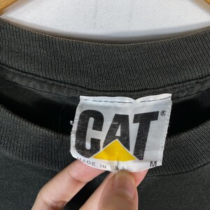 May include: Close-up of a dark gray t-shirt with a white tag. The tag features the word "CAT" in bold, black letters above a yellow triangle. The tag also reads "MADE IN USA" and the size "M".